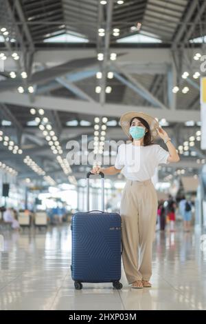 Junge Frau trägt Gesichtsmaske mit Gepäck zu Fuß im Flughafen, Schutz Coronavirus-Krankheit Infektion, asiatische Frau Reisende mit Hut. Zeit zum Trave Stockfoto