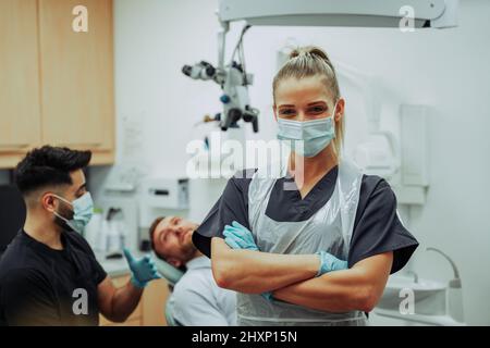 Kaukasische weibliche Zahnärztin, die eine chirurgische Maske trägt, die mit gekreuzten Armen im Ärztezimmer steht Stockfoto
