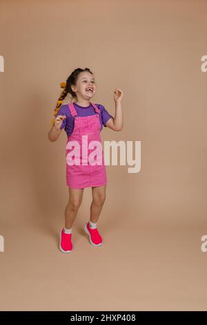 Entzückt begeistert hüpfendes kleines Kind mit gelben Kanekalon-Zöpfen, die tanzten und lächelnd mit fehlenden Zähnen in pinkem Jumpsuit, violettem T-Shirt aufschauten Stockfoto