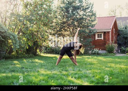 Glückliche übergewichtige Frau, die Yoga-Übungen macht, steht auf dem Boden und dehnt den Körper auf dem Hinterhof der Hütte mit Holzhaus und Bäumen im Hintergrund aus Stockfoto