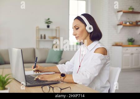 Eine Frau mit Kopfhörern sitzt am Schreibtisch mit Laptop, schaut sich einen Videovortrag an und macht sich Notizen Stockfoto