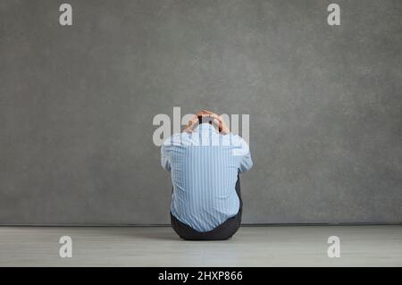 Rückansicht eines Mannes, der auf dem Boden sitzt, den Kopf hält, sich bekümmert, frustriert und hoffnungslos fühlt Stockfoto