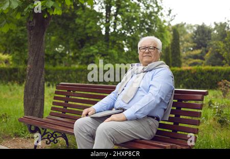 Porträt eines glücklichen älteren Mannes, der einen Laptop in der Hand hält und auf einer Bank im grünen Sommerpark sitzt Stockfoto