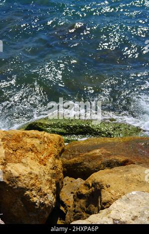 An einem sonnigen Tag treffen kleine Wellen auf nasse Felsen am Meer Stockfoto