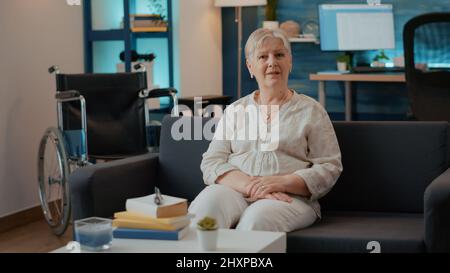 Porträt einer behinderten Frau im Ruhestand, die auf dem Sofa sitzt, neben dem Rollstuhl im Wohnzimmer, um bei der Mobilität und beim Transport zu helfen. Erwachsene genießen Ruhestand und körperliche Rehabilitation. Stockfoto