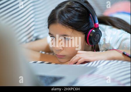 Nahaufnahme eines Mädchens, eines Kindes, das auf dem Sofa liegt und Musik über Kopfhörer hört und einen Laptop benutzt. Stockfoto