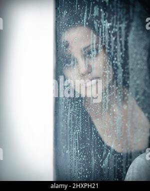 Wenn es mehr ist als nur der Blues. Eine traurige junge Frau, die an einem Fenster sitzt und draußen regnet. Stockfoto