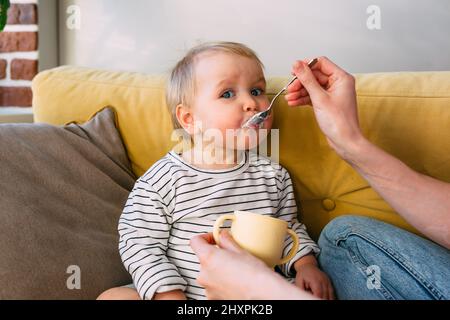 Mama füttert ein kleines Kind zu Hause mit Joghurt aus einem Löffel. Familienkonzept Stockfoto