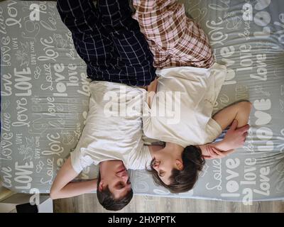 Glückliches Paar, das im Bett in gemütlichem Haus liegt und Kaffee trinkt. Junger Mann in einem T-Shirt und eine niedliche Frau. Familienurlaub und Zweisamkeit, Liebe. Wochenende. Stockfoto