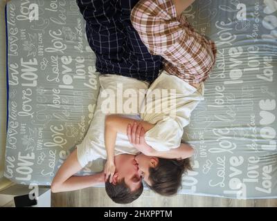 Glückliches Paar, das im Bett in gemütlichem Haus liegt und Kaffee trinkt. Junger Mann in einem T-Shirt und eine niedliche Frau. Familienurlaub und Zweisamkeit, Liebe. Wochenende. Stockfoto