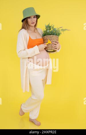 Vertikale moderne zuversichtlich, schwanger lächelnde Frau in grünen Hut, orange Spitze mit Pflanzentopf. Anbau und Pflege, Floristin Stockfoto