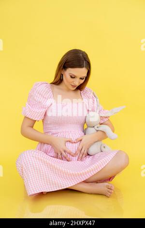 Vertikal froh ruhig ruhig schwanger barfuß Frau sitzend Boden in rosa Kleid mit Haar Kaninchen Spielzeug. Das Mädchen wartet Stockfoto