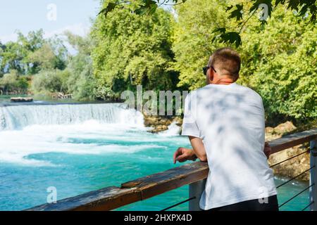 Ein Mann in einem weißen T-Shirt und Shorts steht und schaut an einem sonnigen Tag auf den türkisfarbenen Fluss Stockfoto