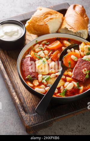 Traditionelle ungarische Babgulyas-Bohnen-Gulaschsuppe aus Pinto-Bohnen, geräuchertem Fleisch, Sellerie, Karotten in der Nähe auf dem Teller auf dem Holztablett. Vertikal Stockfoto
