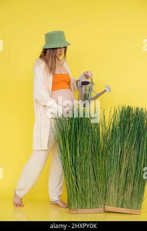 Vertikal von froh schwanger barfuß Frau in hellem Outfit, grüner Hut, hält Bauch mit Gießkanne. Wasserpflanzen, Erde Stockfoto