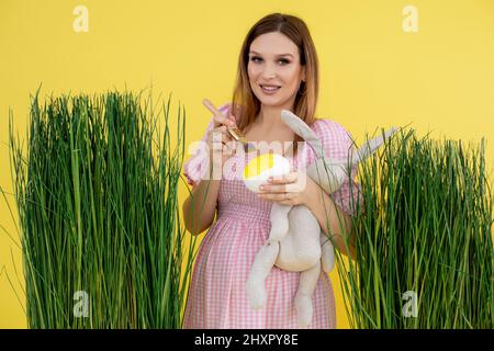 Fröhliche lächelnde Schwangere in hellrosa Kleid in der Nähe von hohem grünen frischen rohen Gras, mit Kaninchen Spielzeug Färbung Osterei Stockfoto