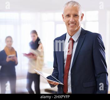 Unser Geschäft ist immer im Kontakt mit der Zeit. Aufnahme eines reifen Geschäftsmannes, der im Vordergrund mit zwei Kollegen hinter ihm steht. Stockfoto