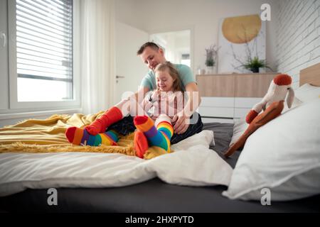 Vater zog seiner kleinen Tochter mit Down-Syndrom verschiedene Socken an, wenn er zu Hause auf dem Bett saß. Stockfoto