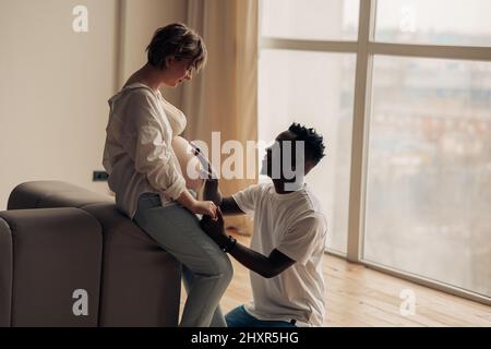 Ein junger glücklicher afrikanischer Mann kniet vor seiner kaukasischen Schwangeren und berührt ihren Bauch an der Hand. Interracial Ehe Konzept. Stockfoto