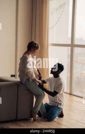 Ein junger glücklicher afrikanischer Mann kniet vor seiner kaukasischen Schwangeren und berührt ihren Bauch an der Hand. Interracial Ehe Konzept. Stockfoto