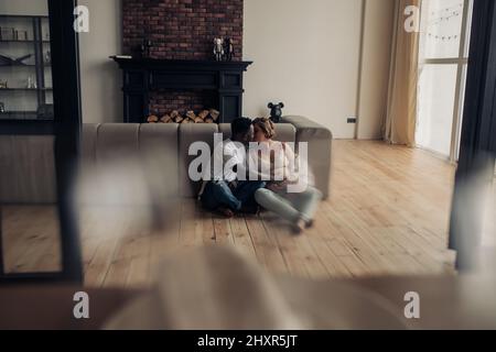Der junge afrikaner und seine kaukasische Schwangere sitzen auf dem Boden neben dem Sofa zusammen und umarmen und küssen sich. Interracial Ehe Konzept. Stockfoto