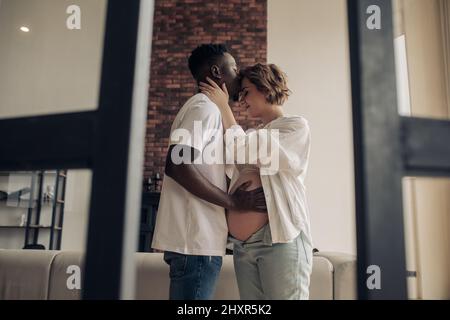 Ein junger glücklicher afrikanischer Mann steht, umarmt, küsst seine kaukasische Schwangere und berührt ihren Bauch an der Hand. Interracial Ehe Konzept. Stockfoto