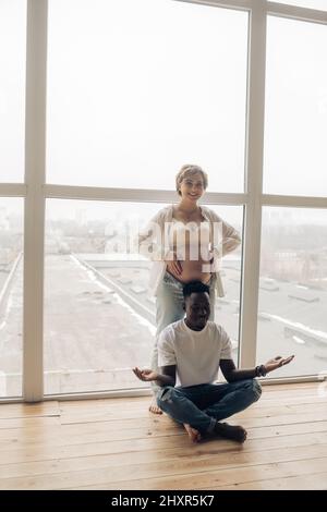 Junger glücklicher afrikanischer Mann meditiert als nächstes seine kaukasische Schwangere gegen das Fenster. Interracial Ehe Konzept. Stockfoto