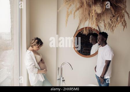 Junge glückliche kaukasische Schwangere steht und berührt ihren Bauch neben einem afrikanischen Mann im Badezimmer vor Fensterhintergrund. Interracial Marriage Konz Stockfoto