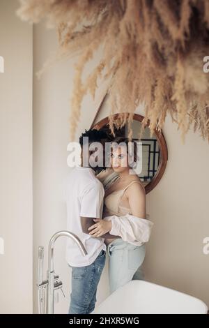 Der junge afrikanische Mann umarmt seine kaukasische Schwangere im Badezimmer gegen Blumenschmuck. Interracial Ehe Konzept. Stockfoto