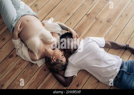 Der junge glückliche afrikanische Mann und seine kaukasische Schwangere liegen auf einem Holzboden und umarmen sich. Interracial Ehe Konzept. Draufsicht. Stockfoto