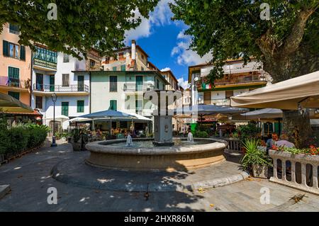 Brunnen und Restaurants im Freien unter den Bäumen auf einem kleinen Platz in Dolceacqua, Italien. Stockfoto