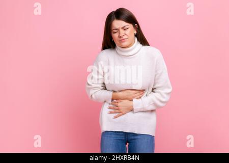 Kranke Brünette Frau mit akuten Schmerzen, Krämpfen durch Verdauungsstörungen oder Gastritis, Appendizitis-Symptome, trägt weißen Pullover im lässigen Stil. Innenaufnahme des Studios isoliert auf rosa Hintergrund. Stockfoto