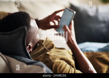 Lassen Sie mich hier ein paar Gespräche führen. Eine kurze Aufnahme eines nicht erkennbaren Mannes, der ein Smartphone benutzt, während er sich zu Hause auf seiner Couch entspannt. Stockfoto