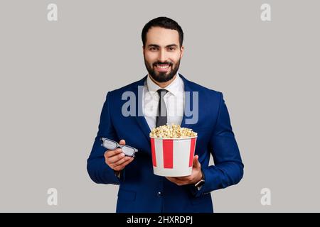 Lächelnder bärtiger Geschäftsmann, der 3D Gläser und Popcorn in der Hand hält, bereit ist, sich einen interessanten Film anzusehen und einen offiziellen Anzug trägt. Innenaufnahme des Studios isoliert auf grauem Hintergrund. Stockfoto