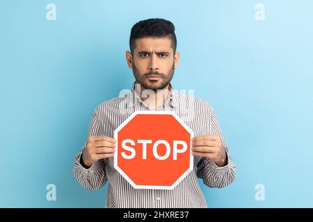 Porträt eines ernsthaften bärtigen Geschäftsmannes, der das Straßenverkehrsschild „Stop“ als Symbol für Verbot, Einschränkungen hält, die Kamera anschaut und ein gestreiftes Hemd trägt. Innenaufnahme des Studios isoliert auf blauem Hintergrund. Stockfoto