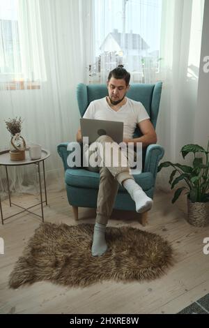Junger Mann, der Laptop-Schreibarbeiten verwendet und zu Hause auf dem Sessel sitzt Stockfoto