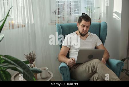 Junger Mann, der Laptop-Schreibarbeiten verwendet und zu Hause auf dem Sessel sitzt Stockfoto