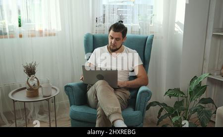 Junger Mann, der Laptop-Schreibarbeiten verwendet und zu Hause auf dem Sessel sitzt Stockfoto