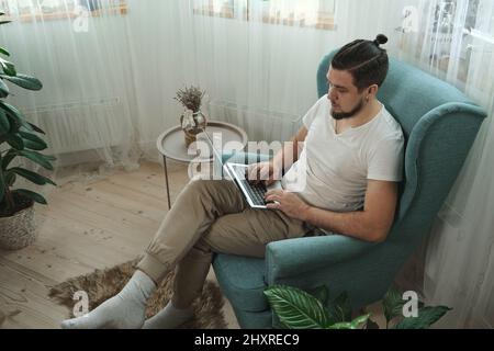 Junger Mann, der Laptop-Schreibarbeiten verwendet und zu Hause auf dem Sessel sitzt Stockfoto