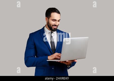 Lächelnder bärtiger Mann, der einen Laptop hält, auf der Tastatur tippt, im Internet surft, Freiberufler, der am Computer arbeitet und einen offiziellen Anzug trägt. Innenaufnahme des Studios isoliert auf grauem Hintergrund. Stockfoto