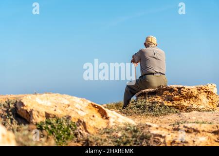 Nachdenklicher alter Mann in Baskenmütze sitzend und mit Gehstock am Ufer am Meer in Spanien Stockfoto