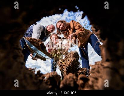 Weltumwelttag der Wiederaufforstung als Konzept der Ökologisierung von Natur und Ökologie. Glückliche Familie mit doggie mit Schaufel zum Graben und Pflanzen Sämlinge Baum wächst in Boden. Blick von der Erdgrube Stockfoto