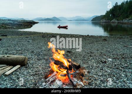 Ein Lagerfeuer an einem felsigen Strand mit Bergen in der Ferne Stockfoto