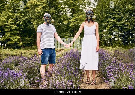 Junges Paar, das in einem Lavendelfeld steht und Gasmasken trägt und die Hände hält. Menschen mit Allergien und blühenden Lavendelblüten. Konzept der saisonalen Allergie Stockfoto