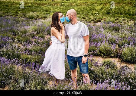 Junges Paar, das im Lavendelfeld steht und Gesichtsmasken trägt. Frau in weißem Kleid, Mann in Shorts und T-Shirt, der sich anschaut. Romantik in Beziehungen, Allergiekonzept Stockfoto