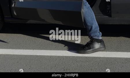 Mann, der auf der Fahrerseite ins Auto fährt. HDR. Nahaufnahme von männlichen Beinen in schwarzen Schuhen und Jeans auf dem Hintergrund von Asphaltstraße und PKW. Stockfoto