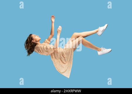 Schweben in der Luft. Entspanntes Mädchen in gelbem Kleid schweben, Blick nach oben, während fliegen in der Luft, mit komfortablen friedlichen Traum. In voller Länge Studio Schuss isoliert auf blauem Hintergrund, innen Stockfoto