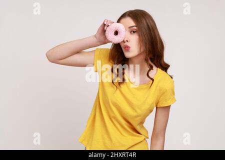 Portrait von positiven lustigen lockigen Teenager Mädchen im urbanen Stil gelb T-Shirt Blick durch Donut und macht küssen Geste, Spaß zu haben. Innenaufnahme des Studios isoliert auf grauem Hintergrund. Stockfoto