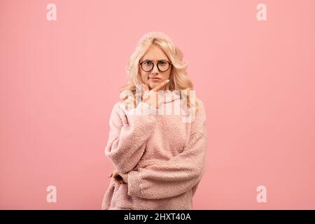 Ernsthafte Frau, die über Fragen nachdenkt, Kinn berührt und auf die Kamera schaut, auf rosa Hintergrund stehend Stockfoto
