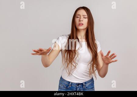 Porträt einer frustrierten Frau mit dunklem Haar, die mit geschlossenen Augen geht, die Hände ausdehnt, um in der Dunkelheit zu suchen, Sichtprobleme, weißes T-Shirt trägt. Innenaufnahme des Studios isoliert auf grauem Hintergrund. Stockfoto